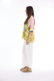 A woman stands in profile wearing a colorful tropical print short-sleeve top with pink sleeves, white wide-leg pants, and tan platform sandals against a plain white studio background.