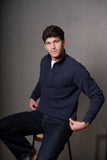 Man wearing a navy blue sweater and jeans sitting on a stool against a gray background