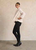 A woman modeling a textured cream-colored cropped jacket with wide sleeves, paired with black pants and black ankle boots, standing against a neutral beige wall.