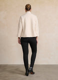 A woman shown from the back wearing a cream-colored textured jacket and black pants, standing against a neutral beige wall.