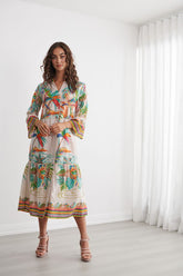 A woman modeling a long-sleeve maxi dress with a colorful tropical palm tree print, standing in a bright, minimalist room with white walls and sheer curtains.