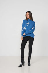 A woman modeling a blue mock-neck knit sweater with white heart patterns, paired with black pants and black ankle boots, against a plain white studio background.