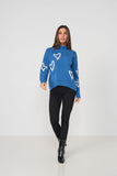 A woman modeling a blue turtleneck sweater with white heart patterns, paired with black pants and black boots, against a plain light gray studio background.