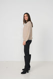 A woman modeling a beige knit sweater, black skinny jeans, and black knee-high boots, standing in profile against a plain white studio background.