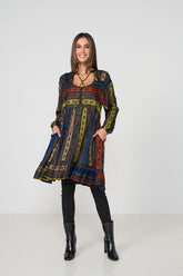 A woman modeling a long-sleeved, V-neck tunic dress with a colorful geometric tribal pattern in blue, yellow, red, and black, paired with black leggings and black ankle boots, against a plain white studio background.