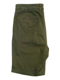 A pair of olive green chino shorts laid flat on a white surface, showing the back pocket with a button closure. The background contains other items and a desk area.