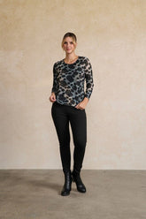 A woman modeling a black and gray leopard-print long-sleeve top, paired with black pants and black ankle boots, standing against a textured beige wall.