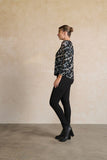 Woman wearing a patterned blouse and black pants against a beige wall.