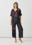 A woman modeling a matching two-piece pajama set with a dark navy blue base and a colorful cosmic print featuring red, pink, and gold planets, stars, and hearts, paired with white strappy high-heeled sandals.