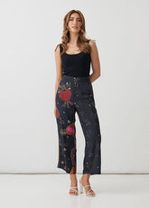 A woman modeling high-waisted cropped pants with a dark navy base and a colorful print of red hearts, planets, and small gold stars, paired with a black ribbed tank top and white strappy heels against a white studio background.