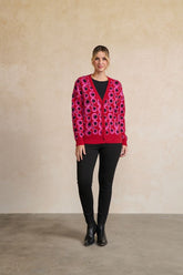 A woman stands against a textured beige wall wearing a bright pink cardigan with a bold black and purple circular pattern, paired with black pants and black ankle boots.