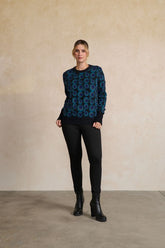 A woman stands in a studio wearing a dark blue and black long-sleeve sweater with a circular geometric pattern, black pants, and black ankle boots against a textured beige wall.