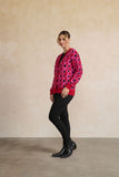 A woman modeling a vibrant pink cardigan with a bold black and purple circular pattern, paired with black pants and black boots, standing against a textured beige wall.