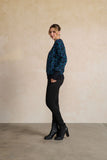 A woman stands in profile wearing a dark blue and black patterned knit sweater, black pants, and black ankle boots against a textured beige wall and concrete floor.