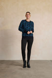 A woman modeling a dark teal and black long-sleeve knit sweater with a circular pattern, paired with black leggings and black ankle boots, standing against a textured beige plaster wall.