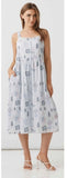 A woman modeling a sleeveless midi dress with a white base and a delicate blue and gray floral and tree print, paired with white strappy high-heeled sandals.