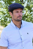 Man wearing a navy cap and light blue shirt with a logo, standing outdoors with greenery in the background