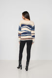 A woman viewed from the back wearing a cream knit sweater with horizontal blue, navy, and brown stripes, paired with black pants and black heeled boots, against a plain light gray studio background.