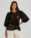 Woman wearing a black lace top with white pants on a plain background