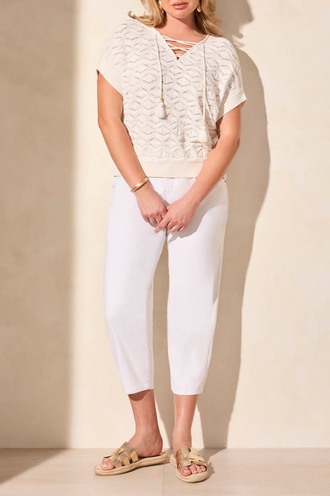 Woman wearing a white lace top and pants with beige sandals on a plain background