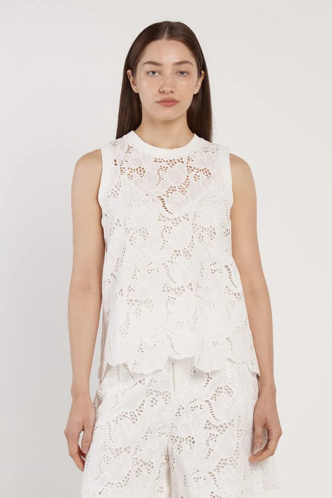 Woman wearing a white sleeveless top with floral lace details on a plain background
