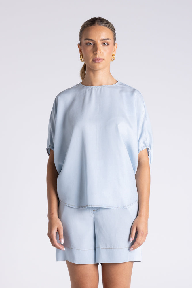 A woman modeling a light blue short-sleeve top and matching shorts against a plain light gray studio background.