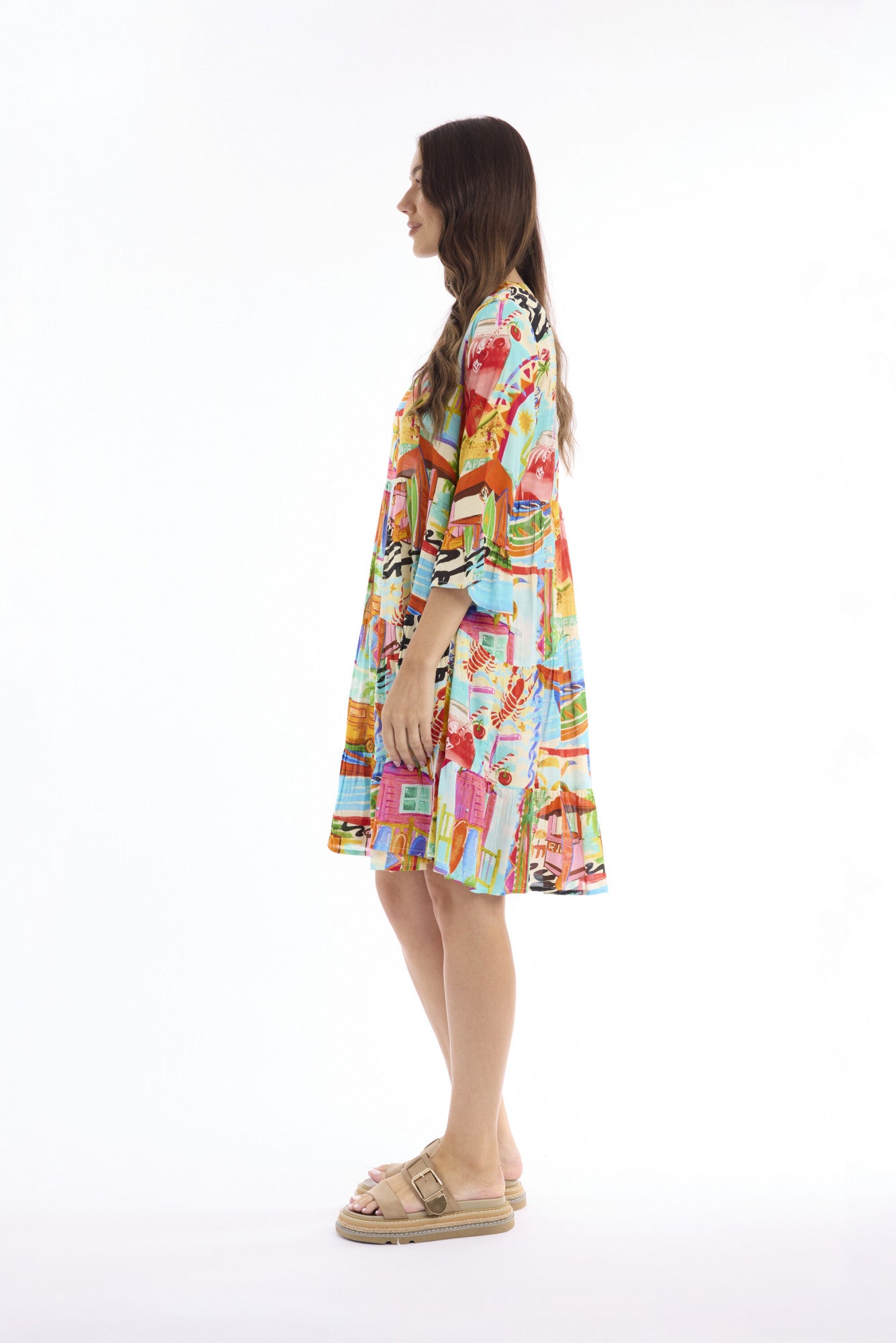 A woman stands in profile wearing a short, knee-length dress with a vibrant multicolor abstract print. The dress features a mix of bright colors including pink, yellow, blue, green, and orange.