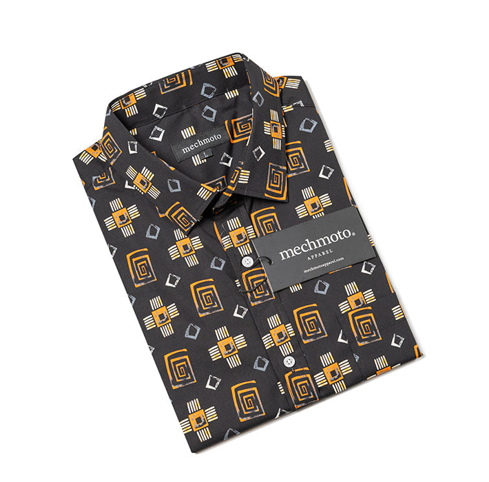 A folded Mechimoto Aztec dress shirt with a black base and a geometric pattern in mustard yellow, white, and gray, shown on a clean white background.