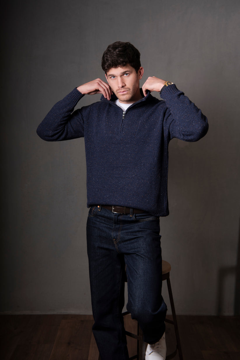 Man wearing a navy blue sweater and dark jeans standing on a stool against a gray background