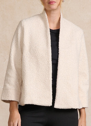 A woman wearing a cropped, open-front textured jacket in a warm off-white or cream bouclé-like fabric, layered over a black top and black pants, against a neutral beige background.