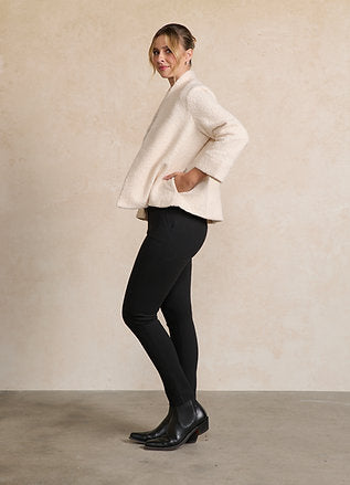 A woman modeling a textured cream-colored cropped jacket with wide sleeves, paired with black pants and black ankle boots, standing against a neutral beige wall.