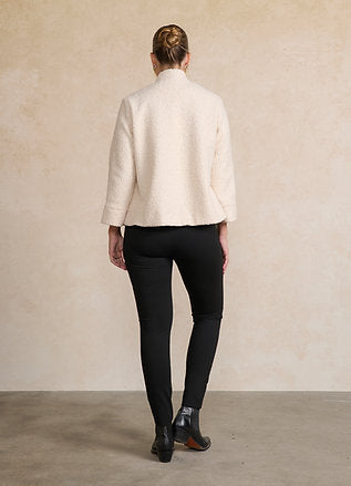 A woman shown from the back wearing a cream-colored textured jacket and black pants, standing against a neutral beige wall.