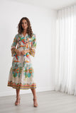 A woman modeling a long-sleeve maxi dress with a colorful tropical palm tree print, standing in a bright, minimalist room with white walls and sheer curtains.