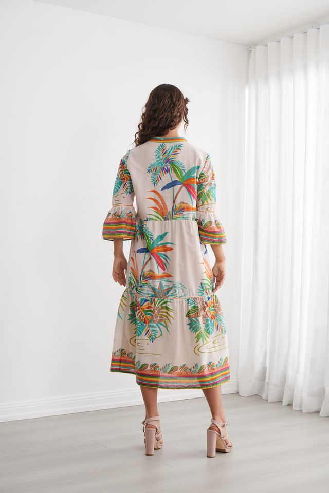 A woman viewed from behind wearing a cream-colored tiered maxi dress with a vibrant tropical print of palm trees and colorful stripes on the sleeves and hem, standing in a bright room with white curtains.