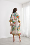 A woman viewed from behind wearing a cream-colored tiered maxi dress with a vibrant tropical print of palm trees and colorful stripes on the sleeves and hem, standing in a bright room with white curtains.