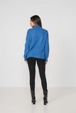 A woman is shown from the back wearing a bright blue mock-neck knit sweater, black skinny pants, and black leather ankle boots, standing against a plain white studio background.