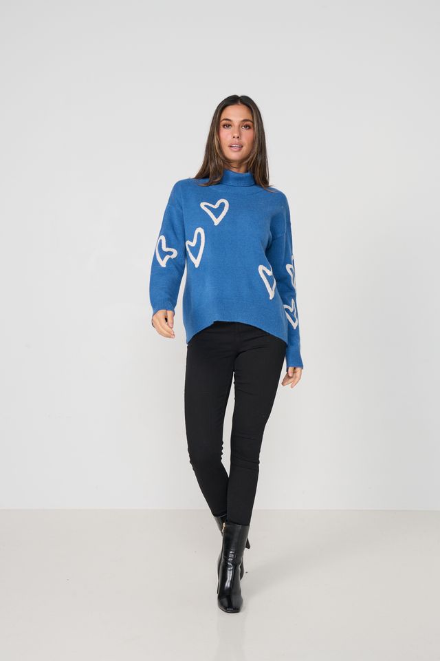 A woman modeling a blue turtleneck sweater with white heart patterns, paired with black pants and black boots, against a plain light gray studio background.