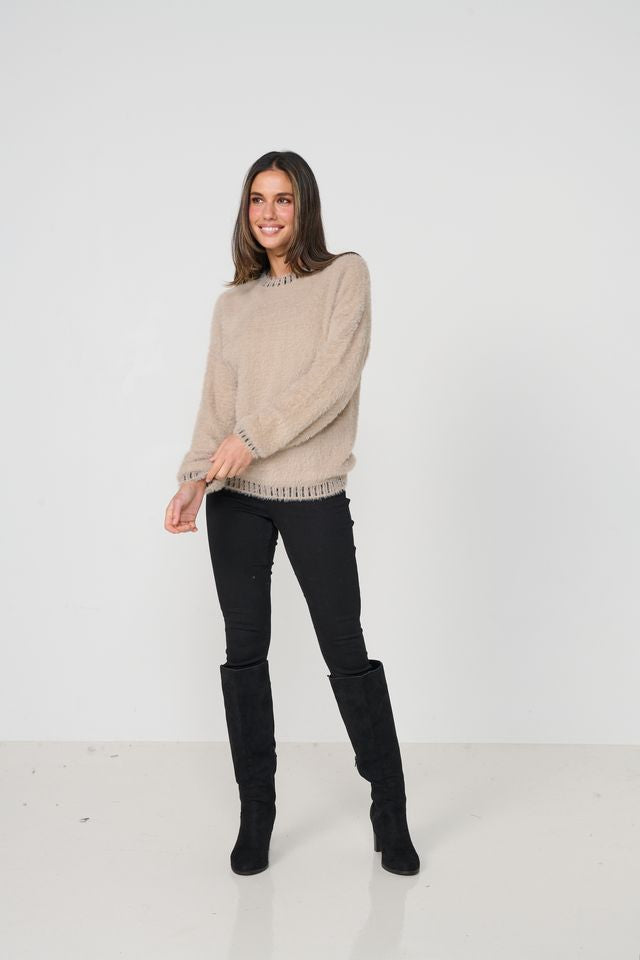 A woman modeling a beige knit sweater with textured fringe details on the cuffs and hem, paired with black skinny jeans and black knee-high boots, against a plain white studio background.
