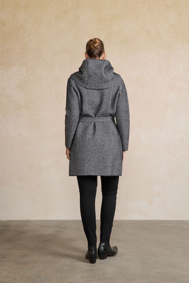 A woman viewed from behind wearing a gray and black patterned hooded coat with a matching belt, paired with black leggings and black boots, standing against a textured beige wall.