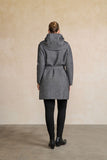 A woman viewed from behind wearing a gray and black patterned hooded coat with a matching belt, paired with black leggings and black boots, standing against a textured beige wall.