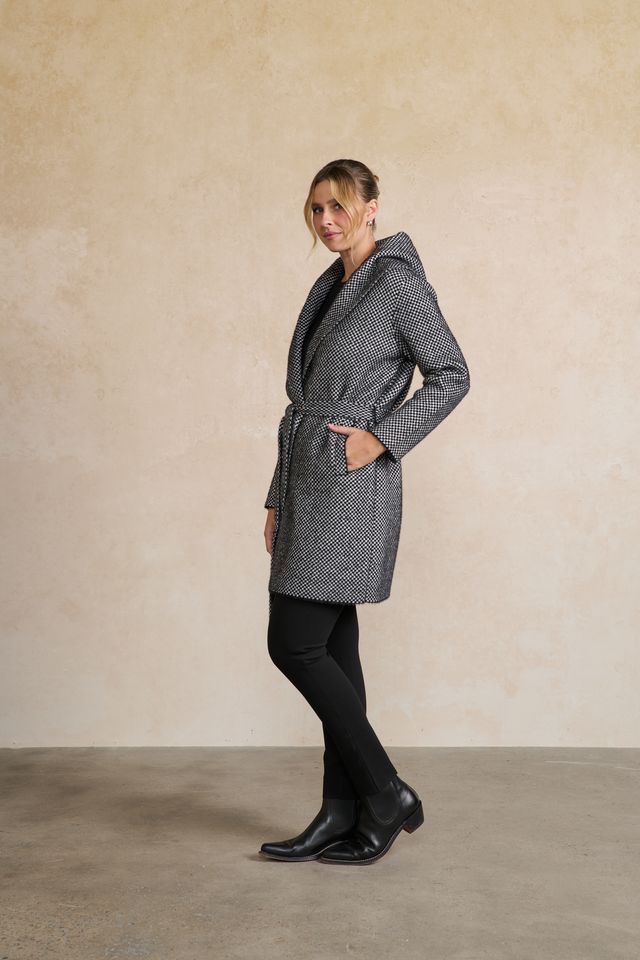 A woman modeling a gray and black micro-patterned hooded coat with a tie belt, paired with black tights and black boots, standing against a textured beige studio wall.