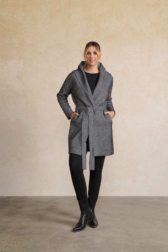 A woman modeling a gray and black micro-patterned wrap coat with a shawl collar and tie belt, paired with black pants and boots, standing against a textured beige studio backdrop.