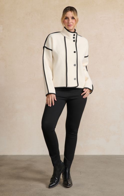 Woman wearing a white jacket with black trim and black pants against a beige background
