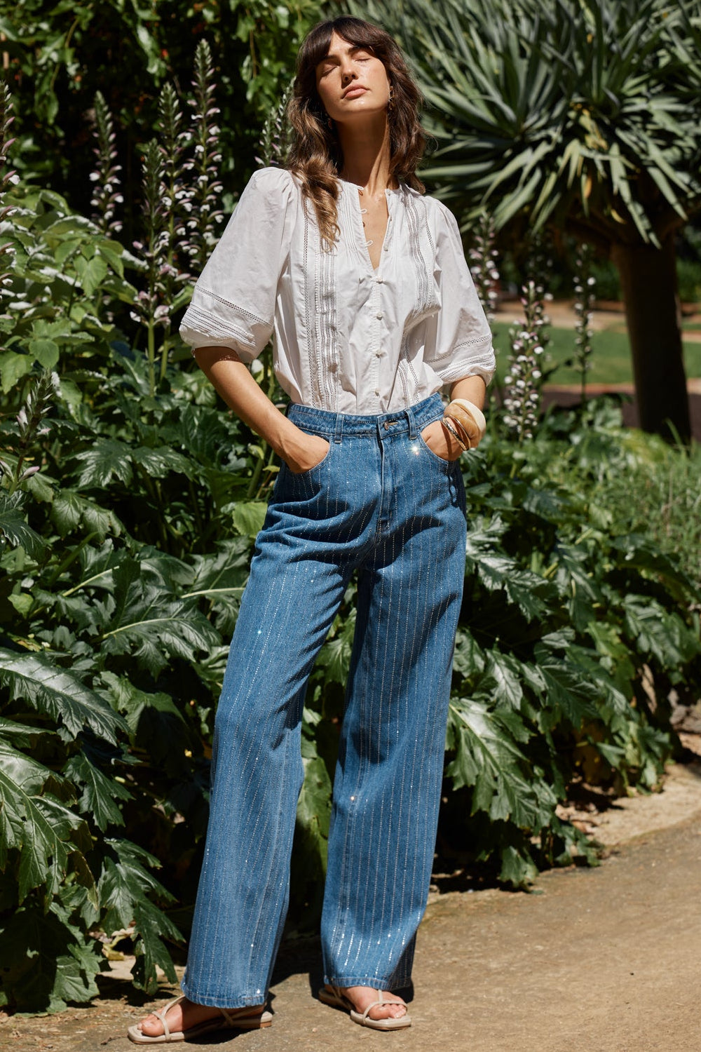 A woman stands outdoors in a garden setting wearing a white long-sleeve button-up blouse and high-waisted, wide-leg blue jeans with thin vertical pinstripes.