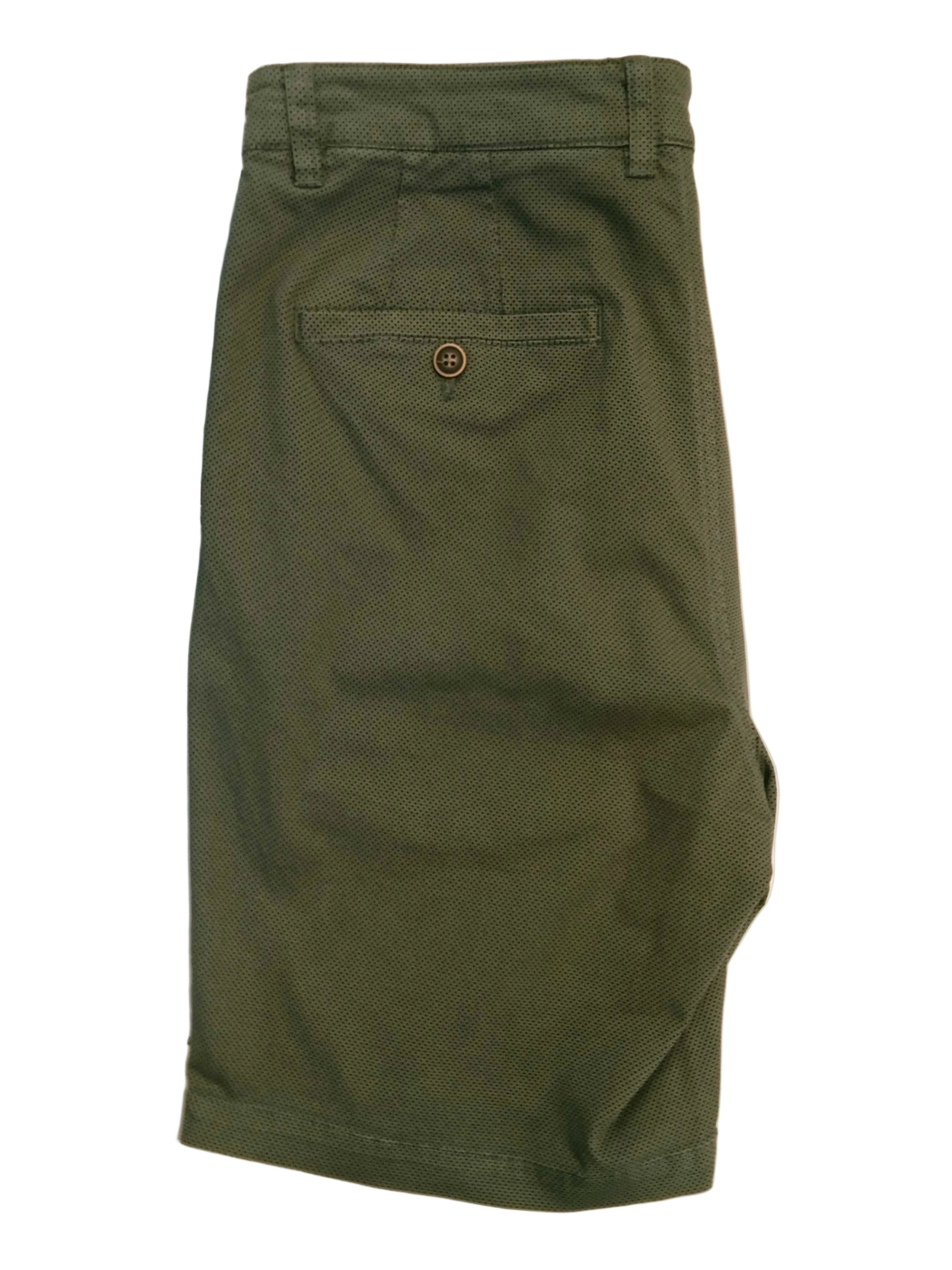 A pair of olive green chino shorts laid flat on a white surface, showing the back pocket with a button closure. The background contains other items and a desk area.