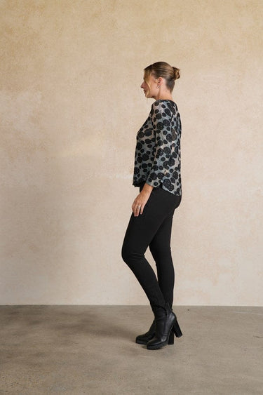 Woman wearing a patterned blouse and black pants against a beige wall.