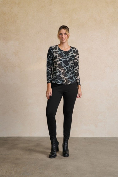 A woman modeling a long-sleeve crewneck top with a black, gray, and white abstract pattern, paired with black pants and black ankle boots, standing against a textured beige wall.