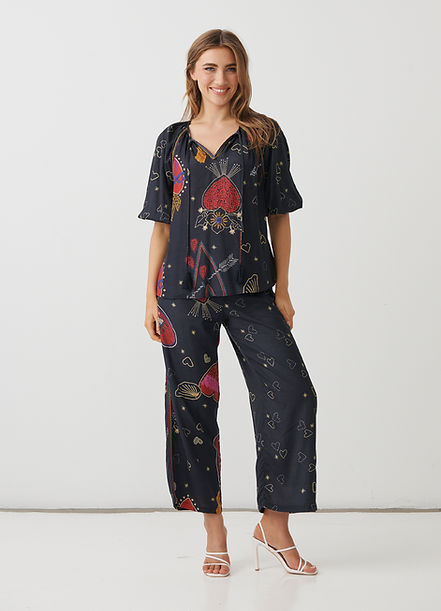 A woman modeling a matching two-piece pajama set with a dark navy blue base and a colorful cosmic print featuring red, pink, and gold planets, stars, and hearts, paired with white strappy high-heeled sandals.