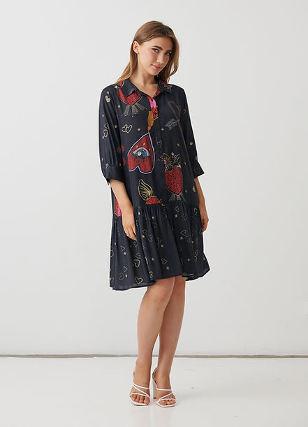 A woman modeling a short black dress with a whimsical red, orange, and gold print featuring strawberries, hearts, and stars, paired with white strappy heels against a plain white studio background.