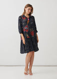 A woman modeling a short black dress with a whimsical red, orange, and gold print featuring strawberries, hearts, and stars, paired with white strappy heels against a plain white studio background.